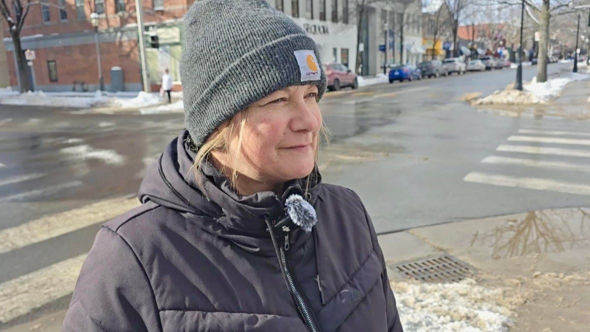 angela-hicks A woman with a grey hat and a winter jacket looks to her left, with a street and snow in the background.