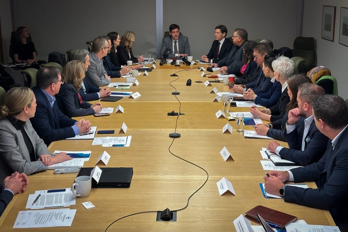 b-c-provincial-task-force-2 A man in a suit sits at the end of a board table with over a dozen professionals all looking at him.