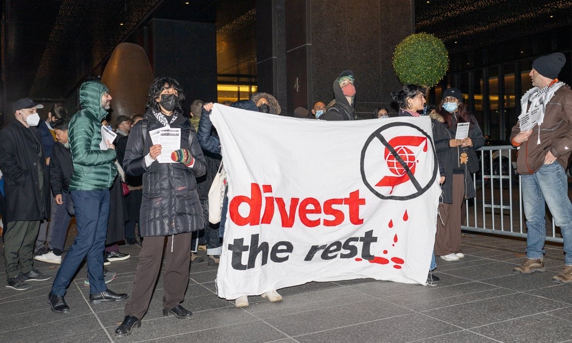 A group of people stand outside, some wearing face masks. Several people are holding a large fabric banner that reads "divest the rest" with a Scotiabank logo crossed out.