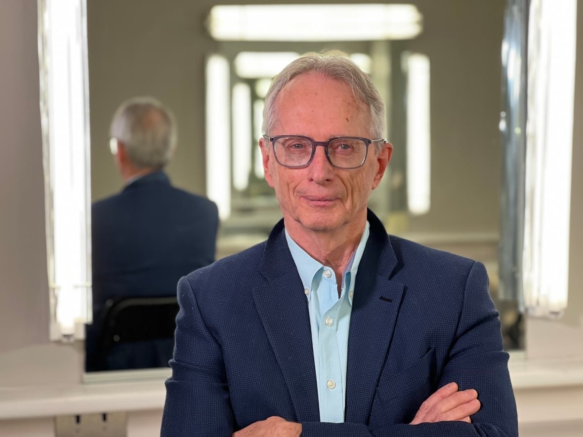 A man in black rimmed glasses stands with arms crossed, with a mirror behind him.
