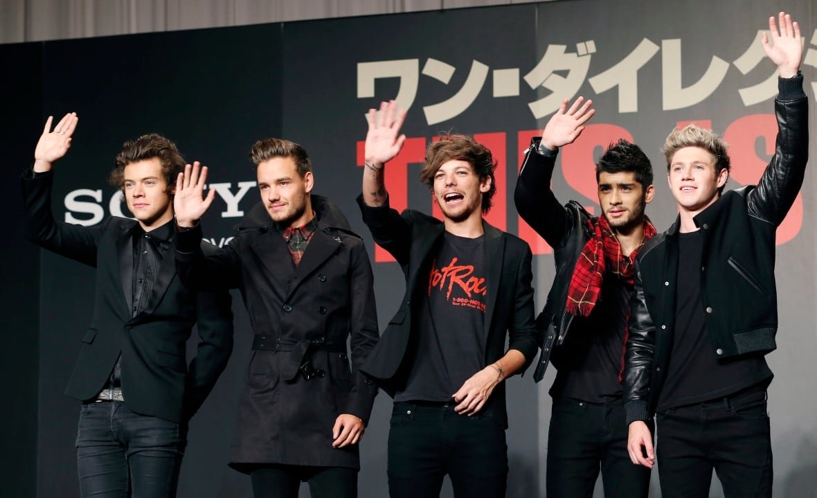 Five young men wave on a red carpet, most wearing all black outfits. There is a black backdrop behind them with Japanese on it.