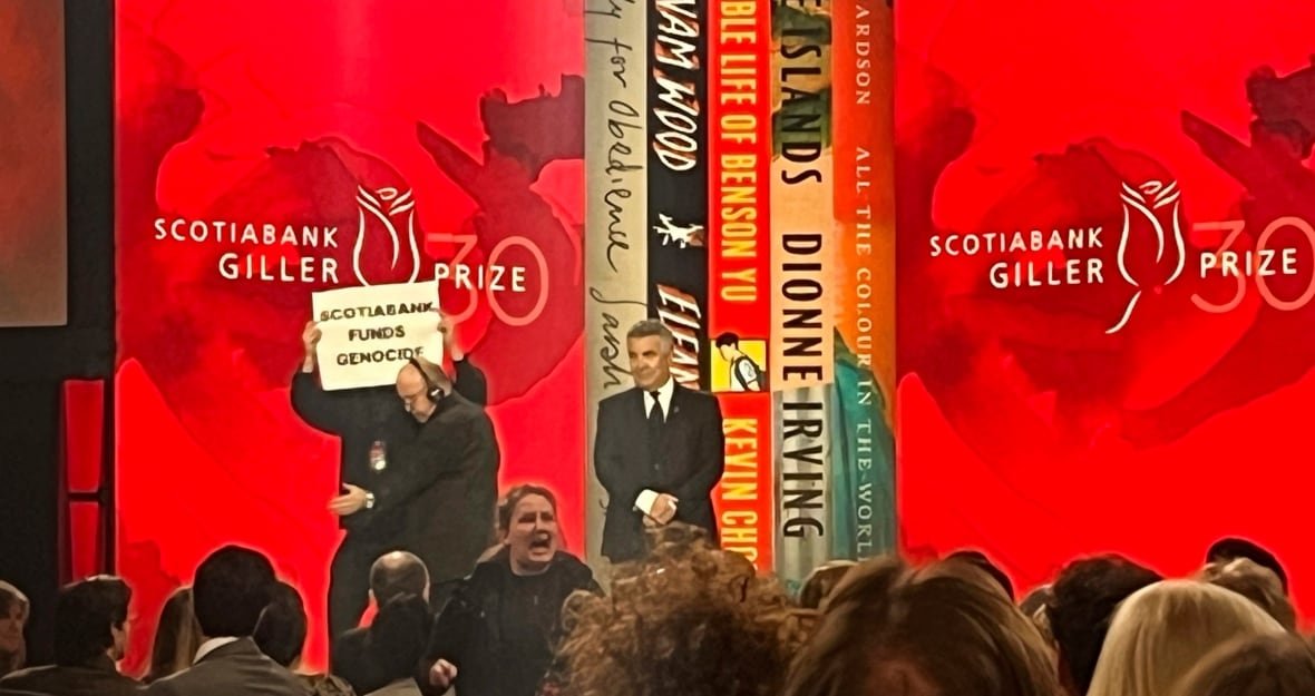 A blurry photo of the stage at the Scotiabank Giller Prize shows security escorting someone offstage as they hold a sign that says 'Scotiabank funds genocide' above their head.