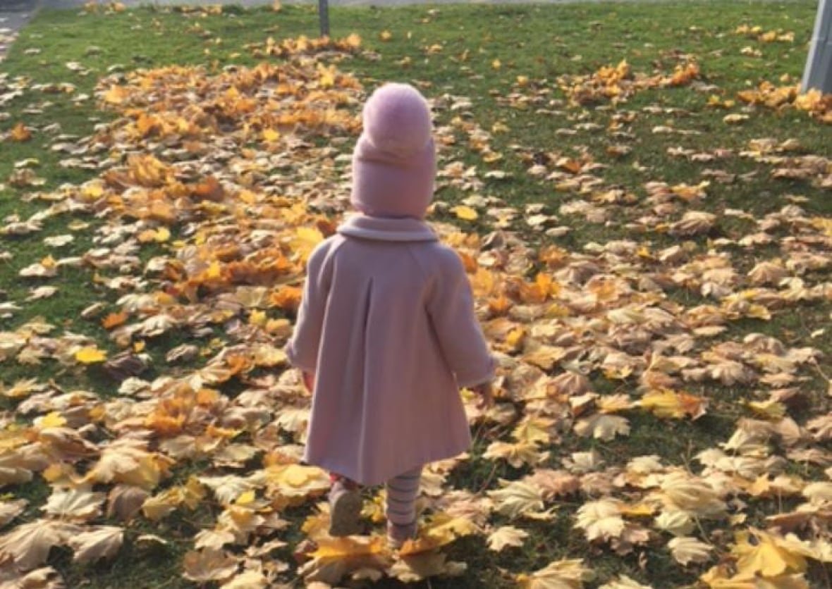 child-in-leaves A little girl dressed in a pink pea coat and pink hat with giant pink pom-pom plays on yellow leaves on green grass. She is facing away from the camera.
