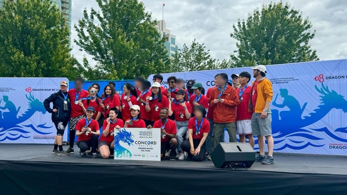 People wearing red shirts and silver medals with blue ribbons stand up on an outdoor stage. Behind them is a banner for a dragon boat race. Some faces have been blurred for privacy.