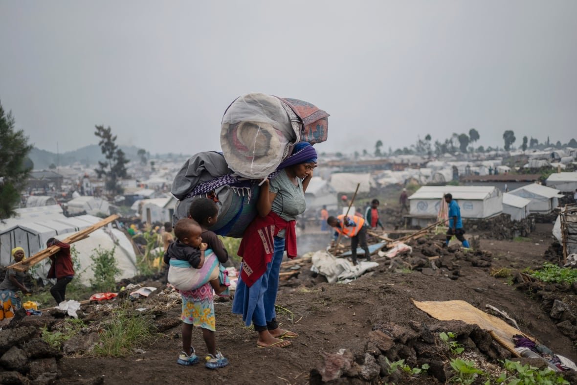 congo-displacement-crisis A woman with her belongings on her back stands near two children.