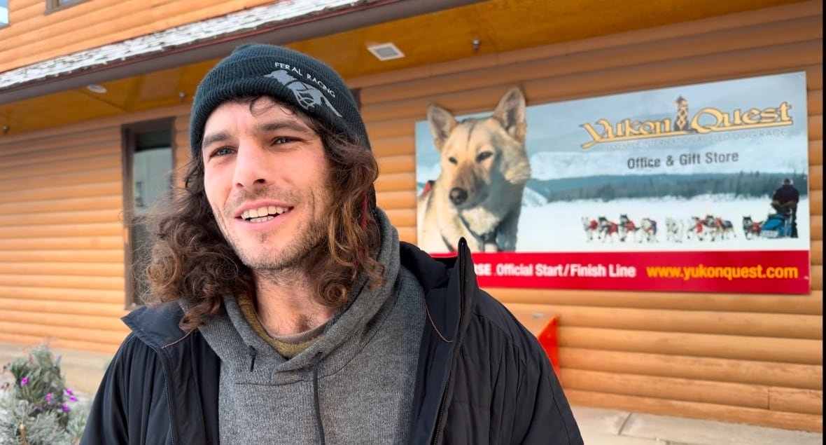 connor-mcmahon A man with long hair and a toque stands in front of a log building.