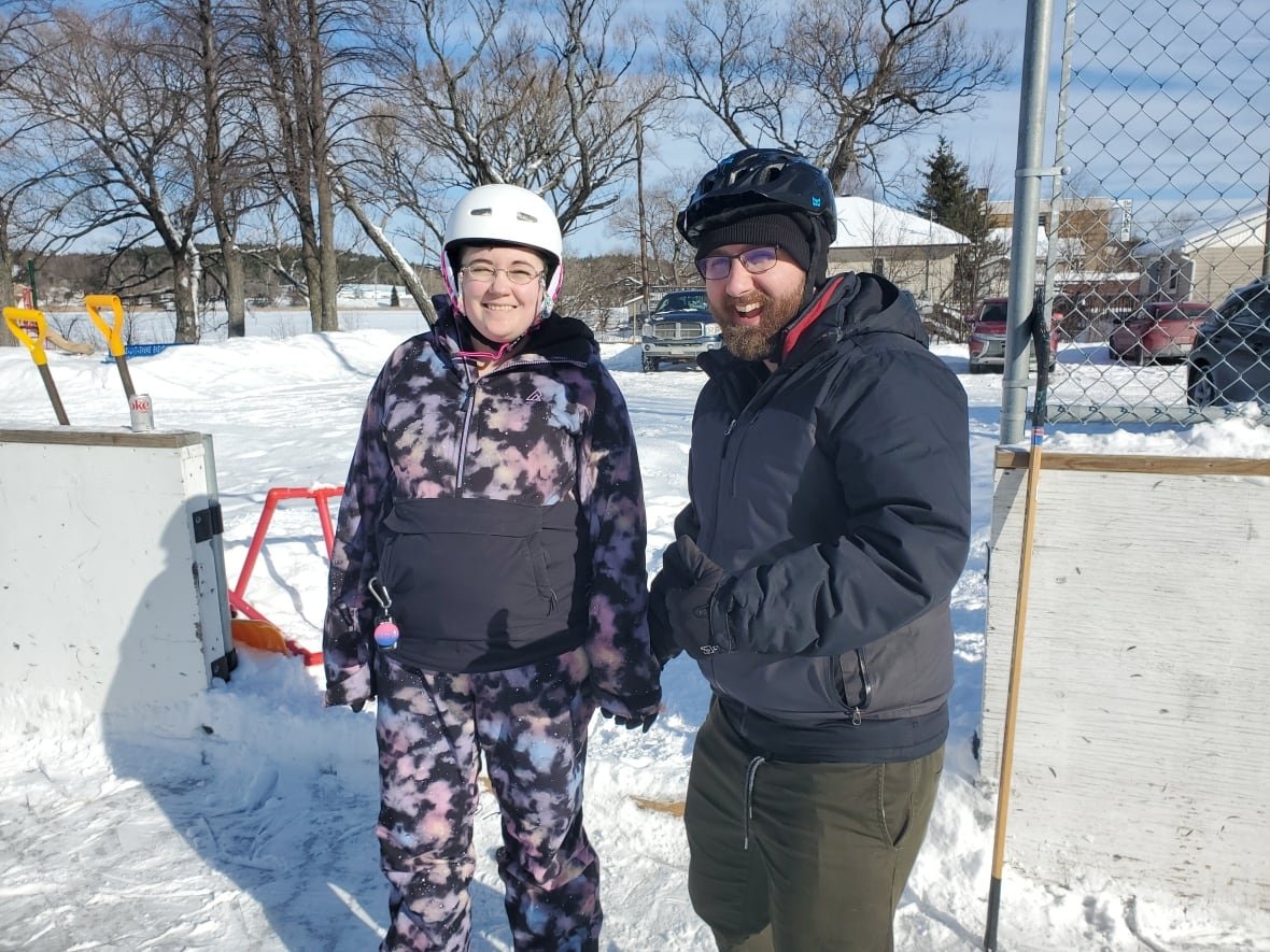 danny-moulelis-and-stefan-scherzinger Two people in snowsuits and helmets standing at the edge of a skating rink.