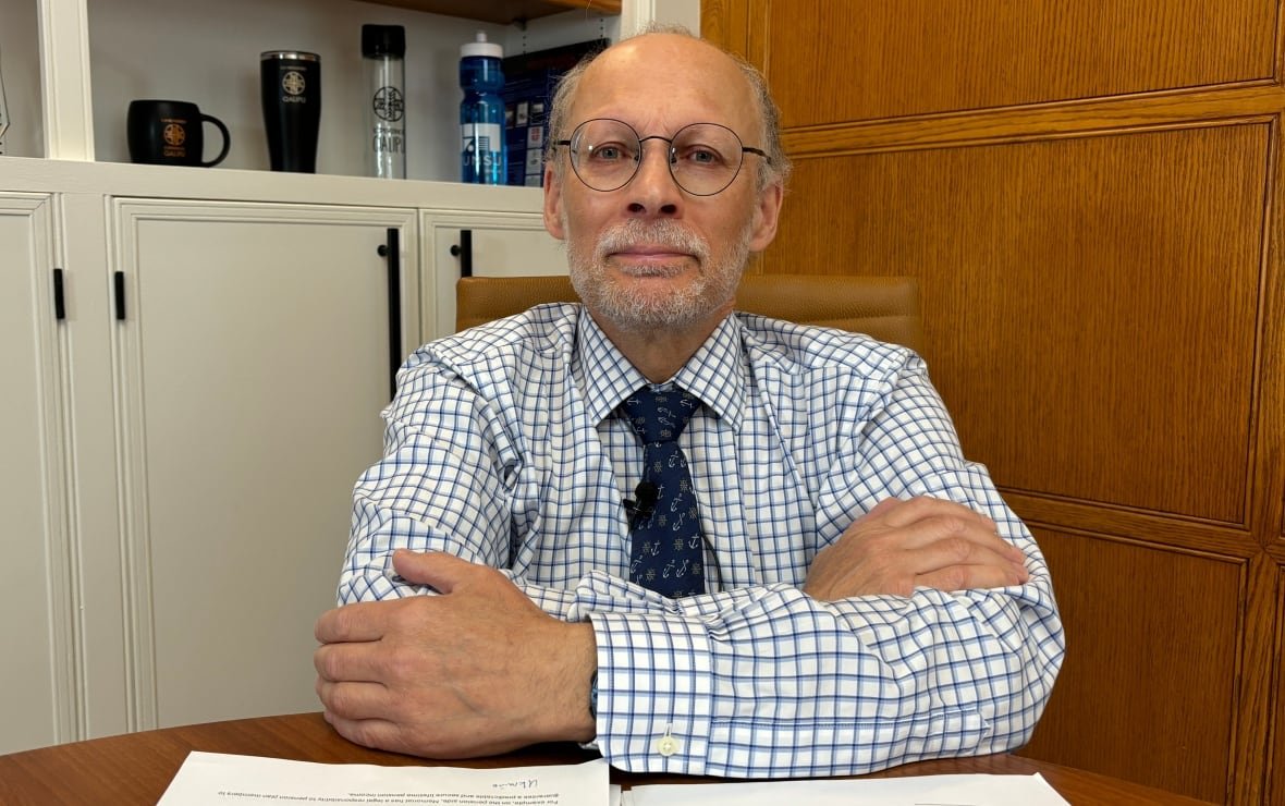 A man with grey hair and rounded glasses has his arms folded on a wooden table in front of him.