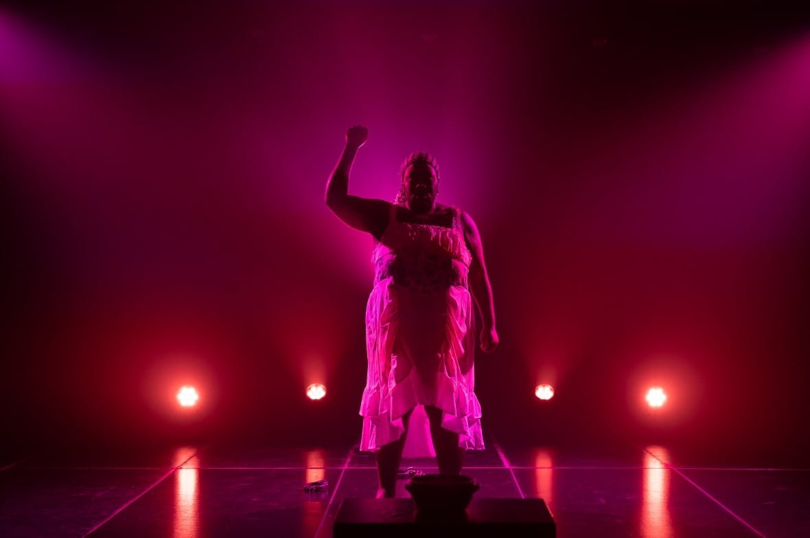 Red and purple lighting light up a stage where a performer in a white dress holds up their right fist in the air.