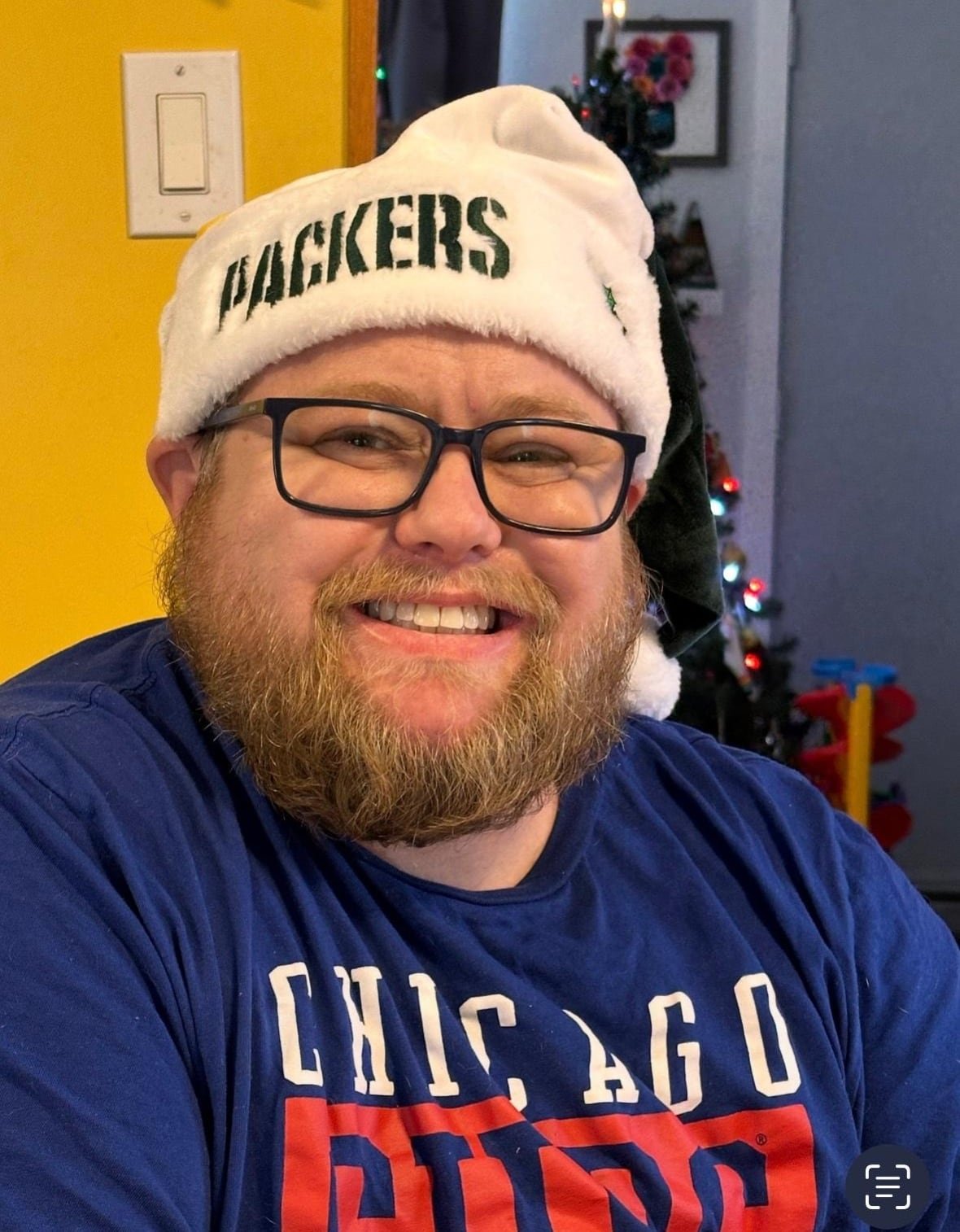 A man with a beard and glasses, wearing a Santa hat with the word 'Packers' on it, smiles for a photo.