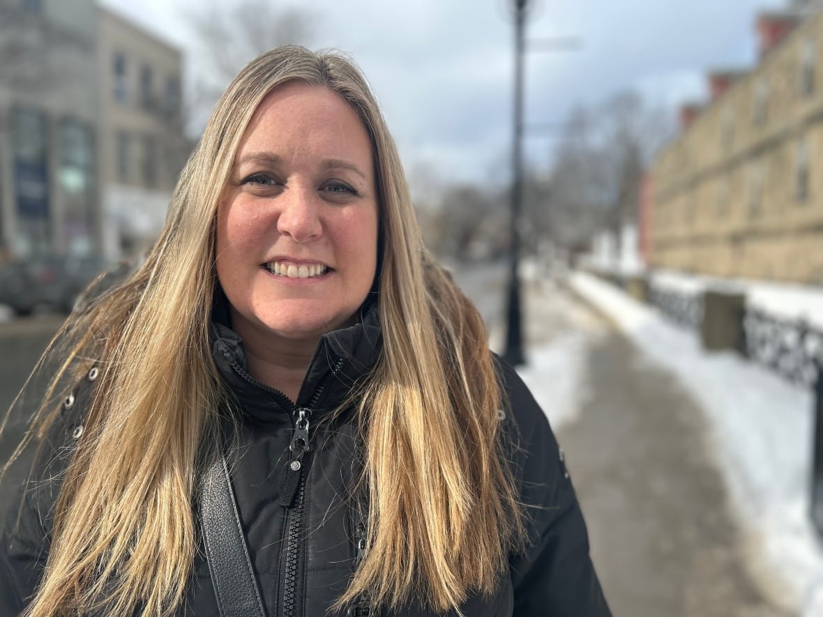 erin-lanigan A woman with long blonde hair smiles at the camera, with snow on the sidewalk behind her.