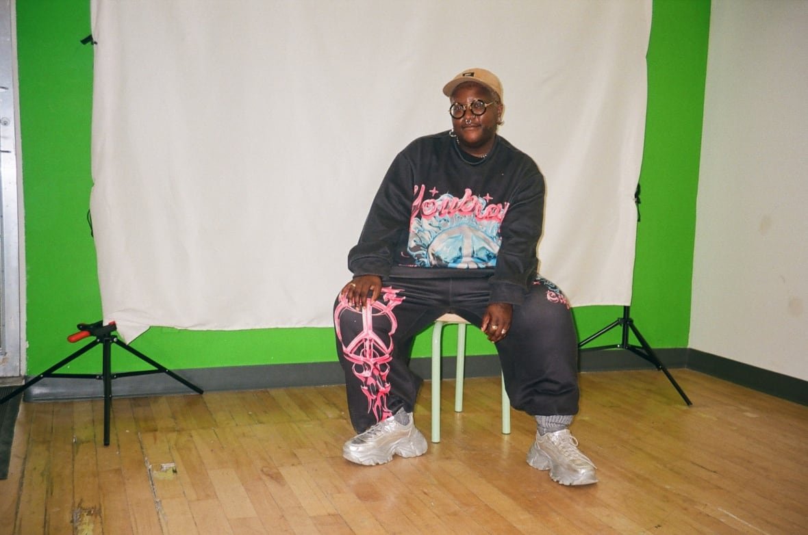 A Black person wearing a black and pink tracksuit with silver running shoes sits in front of a white screen in a studio.