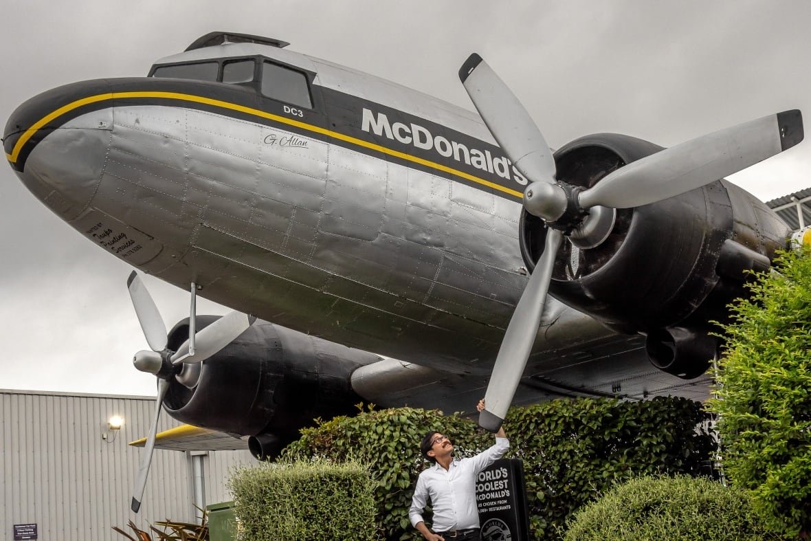 gary-he A mustachioed man stands beneath a large silver airplane with the McDonald's logo on it, touching the propeller and looking up.