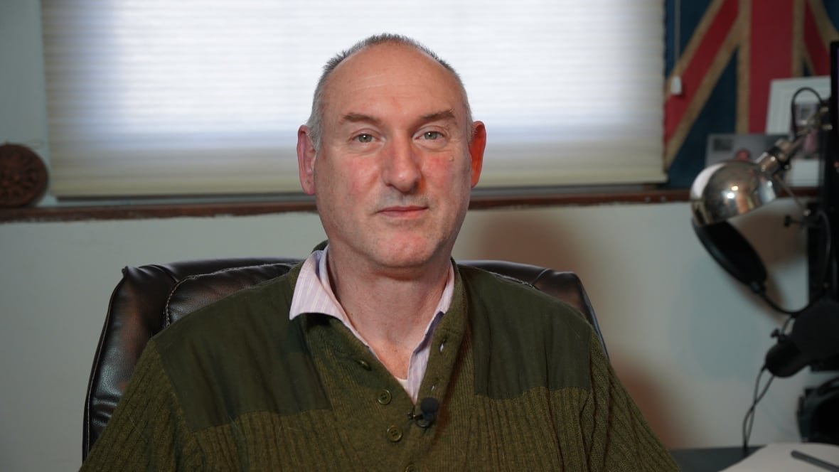 A balding man with close-cropped grey hair wears a green sweater and sits in an office.