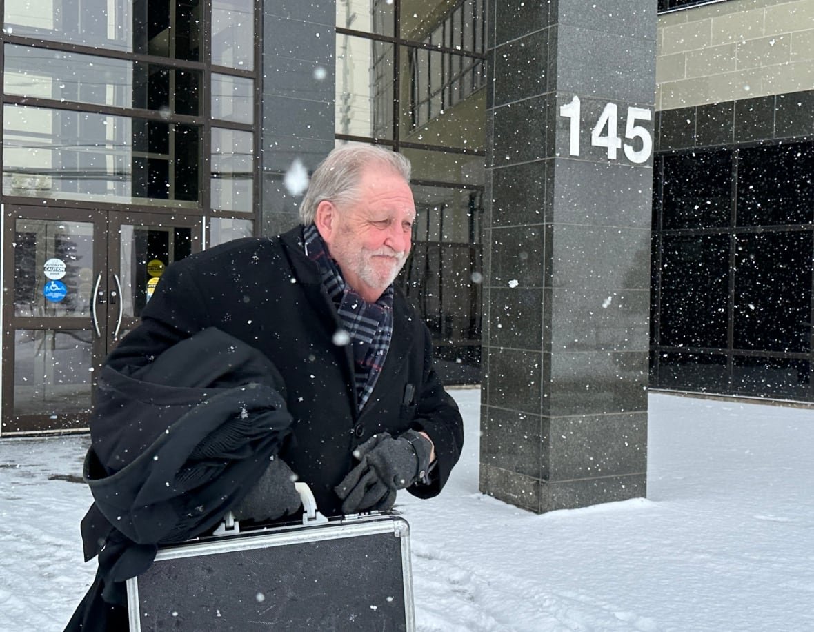 A man in a black coat holding a briefcase walks out of a stone-clad building in the snow.