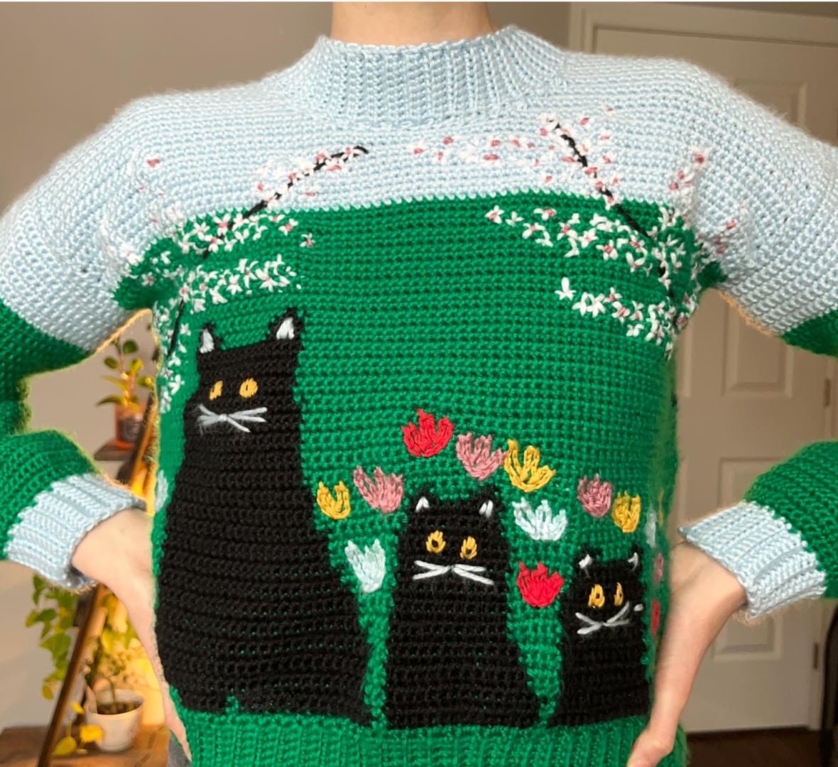 A woman wearing a sweater that depicts a Maud Lewis painting, three black cats on a green background.