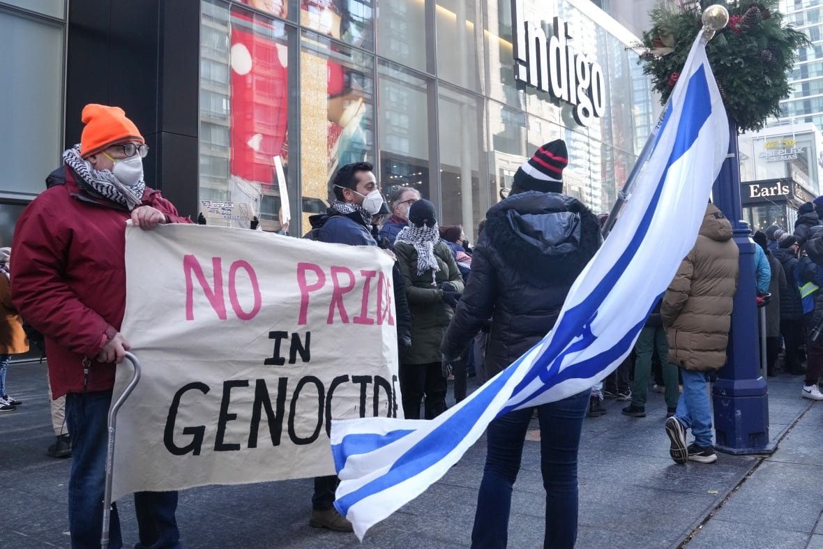 People stand outside of an Indigo bookshop. One is carrying an Israel flag and another two hold a sign that says "No pride in genocide".