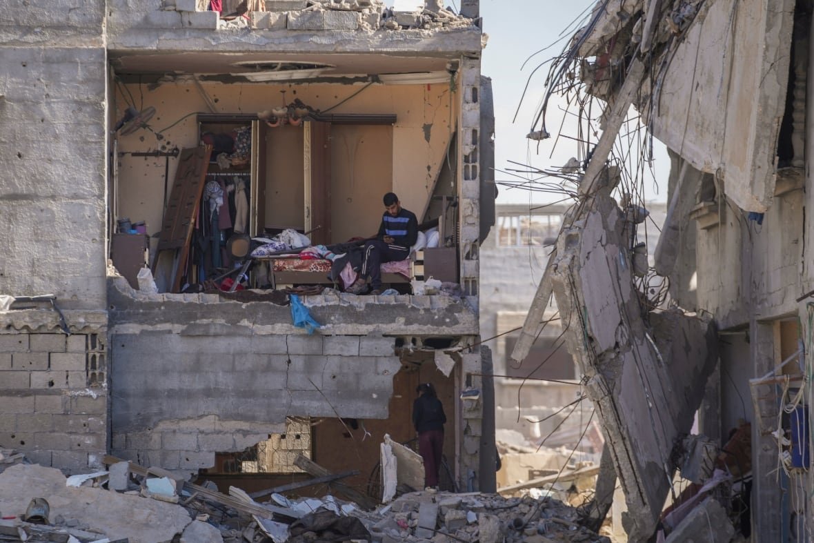 A young man sits in a bedroom of a destroyed building with no wall.