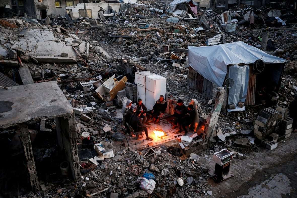 People gather near a fire next to the rubble of destroyed buildings.