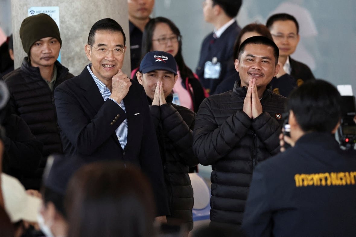 A Southeast Asian man in a dark suit is next to two Southeast Asian men in puffy jackets with their hands pressed together in front of their faces.