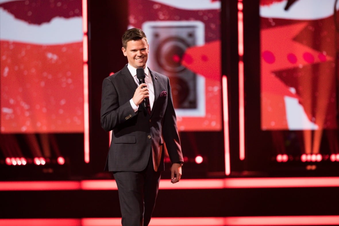 ivan-decker-at-just-for-laughs A man in a suit stands in front of a red background. He has a microphone in his hand and he is smiling