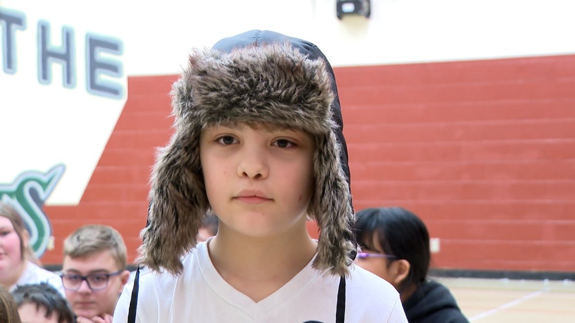 A boy wearing a white t-shirt and Davy Crockett style cap sits on the floor of a gymnasium.