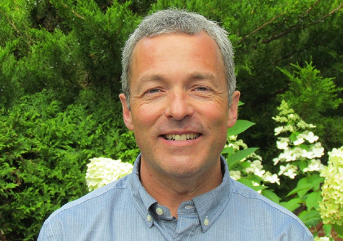 A head and shoulders portrait of a man who looks to be in his forties, with greying hair, wearing a light blue button-down collared shirt and smiling. In the background is greenery and white flowers.
