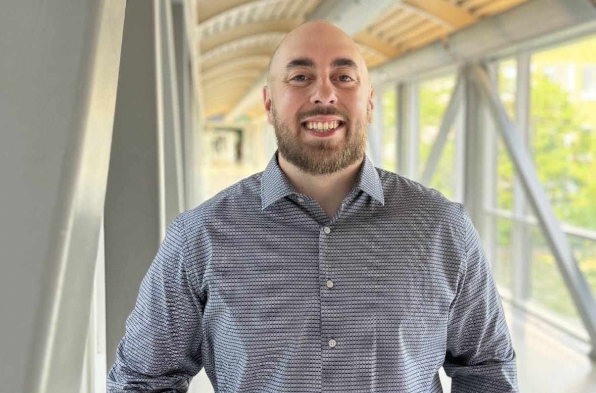 jonathan-hauth A man in a button-down dress shirt smiles as he stands in a bright indoor walkway with large windows looking over a treed area.