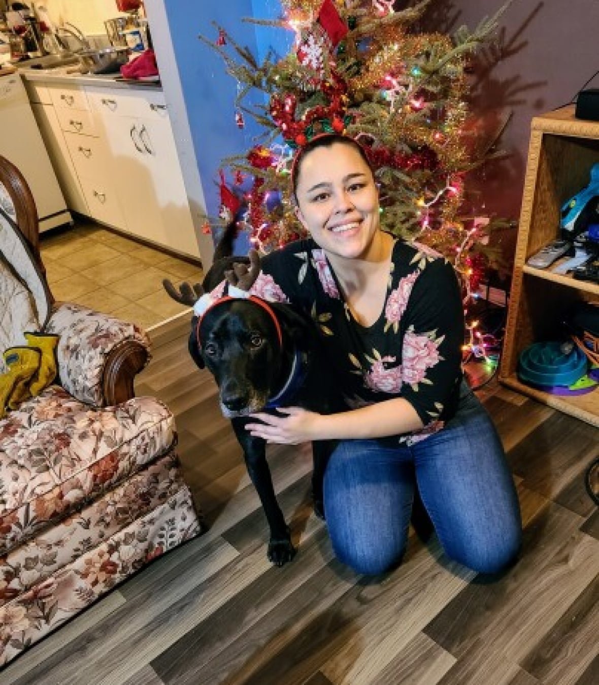 A woman smiles for a photograph while kneeling on the ground next to a black dog.
