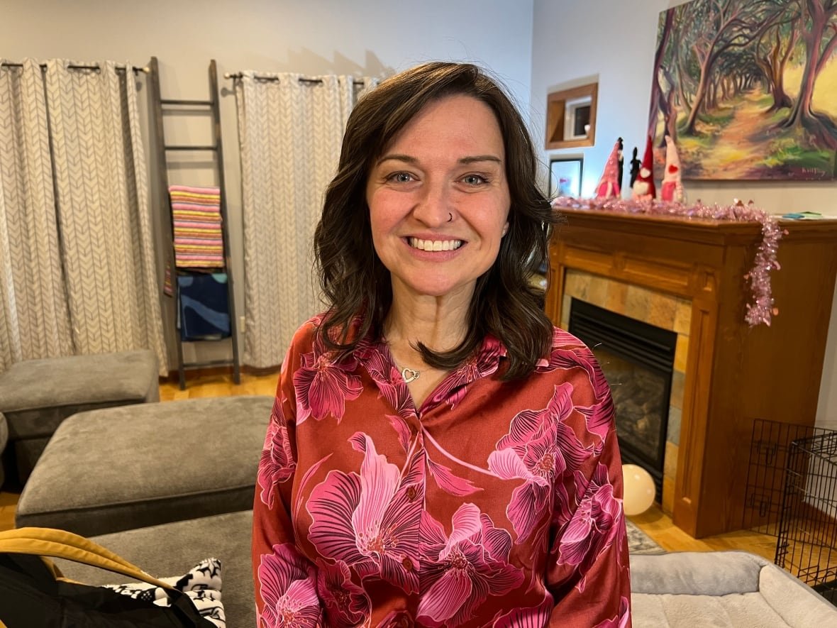 A woman in a pink and red floral blouse poses for a photo while standing in a living room. A fireplace and some couch cushions can be seen behind her.