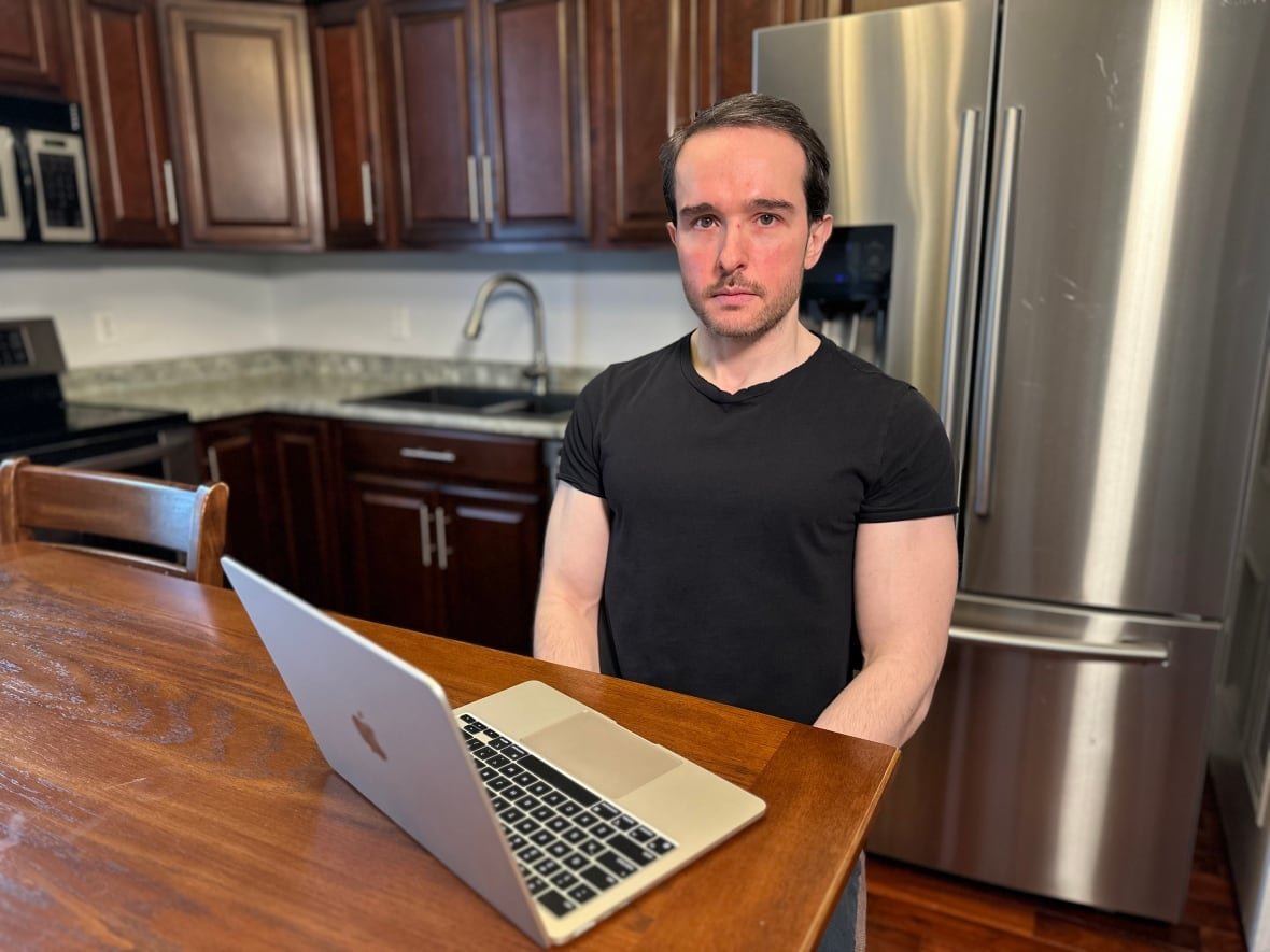 A man stands at a kitchen island with a laptop open in front of him. He's looking at the camera.