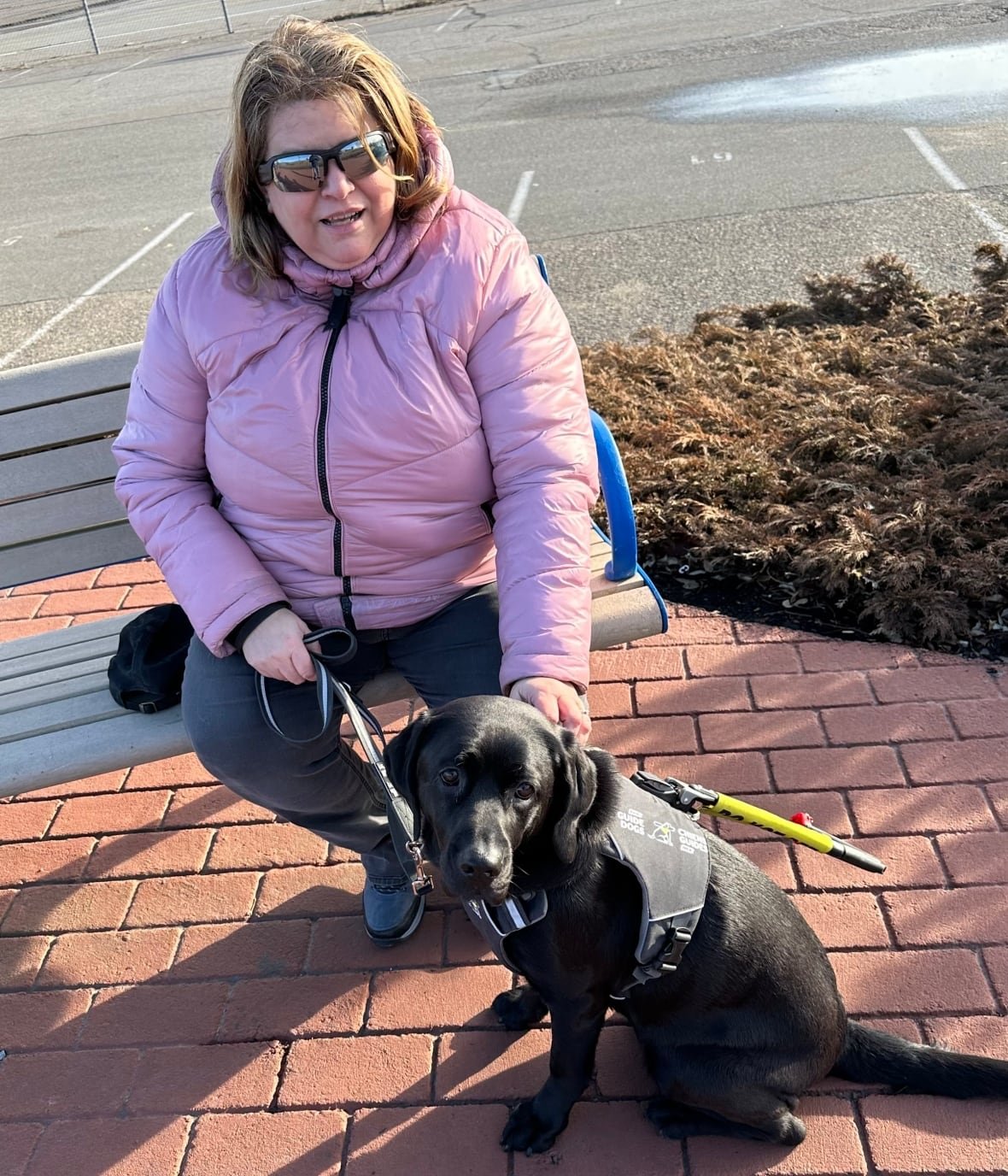 marisa-hersey-misner A woman, wearing sunglasses and a pink jacket, sitting on a park bench. A black guide dog, wearing a harness, sits in front of her.