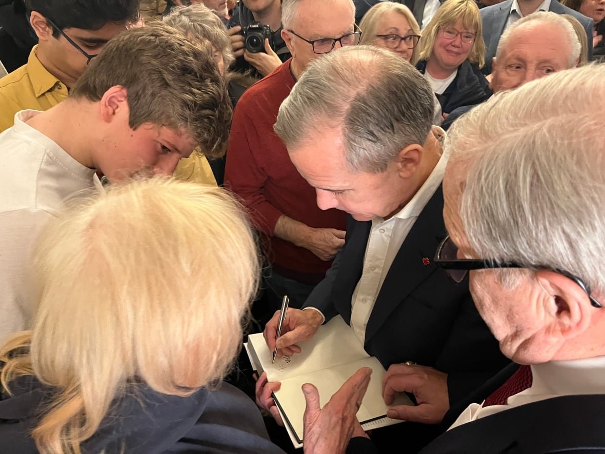 mark-carney-in-pei A man signs an autograph in a book for a woman amid a crowd of people.
