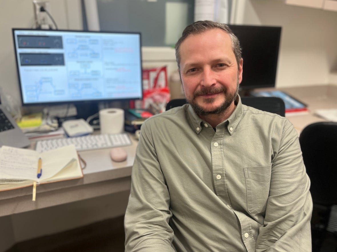A bearded man sitting in front of a computer monitor wearing a light coloured, button-down shirt.