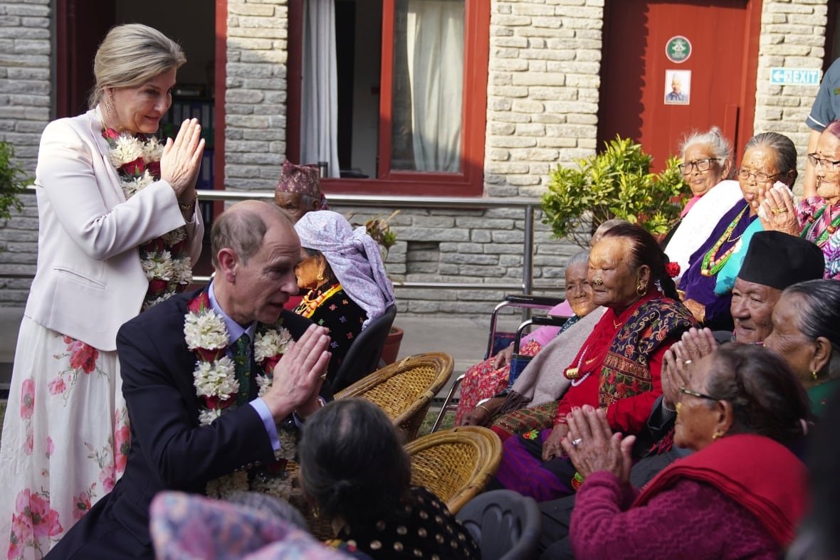 Two people clasping their hands look at several people sitting down, some of whom are also clasping their hands.