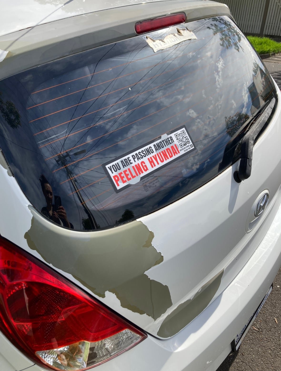 peeling-hyundai The back of a white hatchback car is shown, with peeling paint on the trunk. A sticker in the window reads: You are passing another peeling Hyundai.