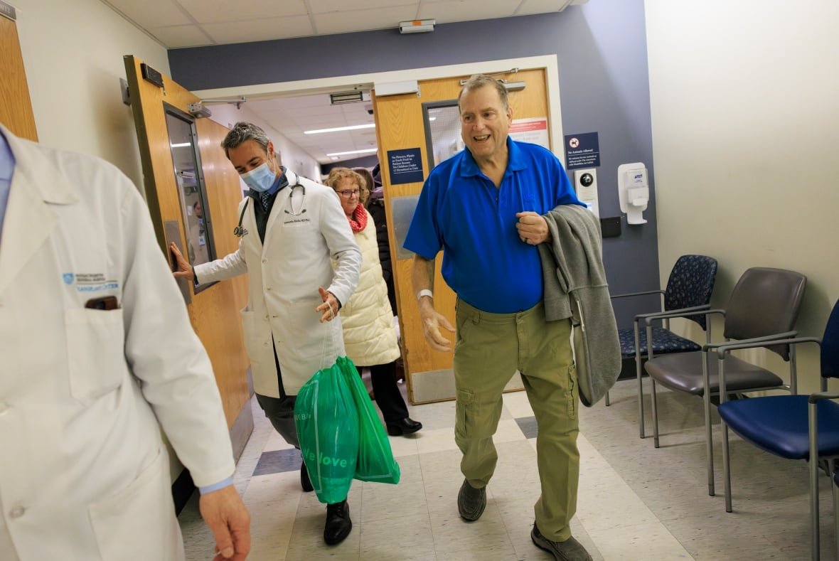 A man in a blue t-shirt with a jacket over his left arm and a hospital band on his right arm walks in a hospital corridor with others.