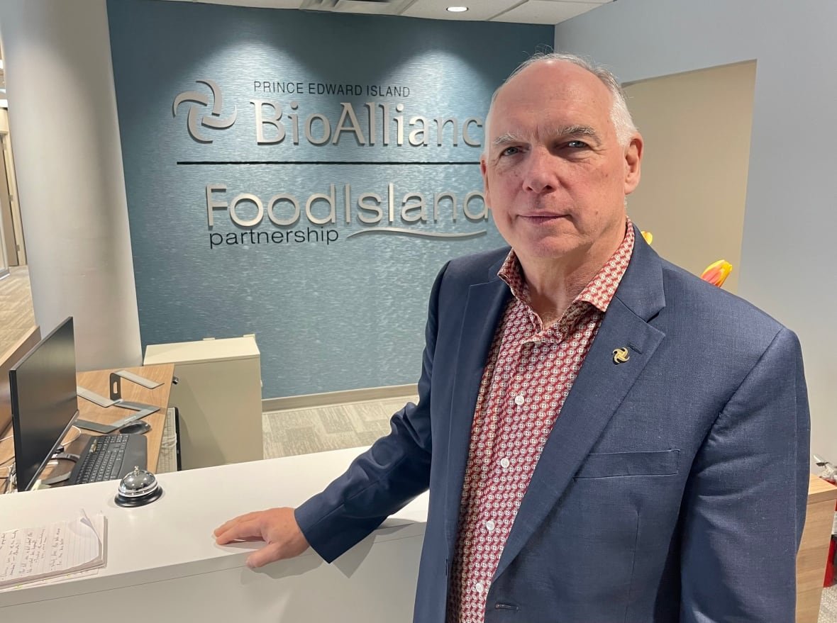 A man in a blue suit jacket and a checked red shirt stands in front of a desk in an office and a sign that reads P.E.I. BioAlliance.