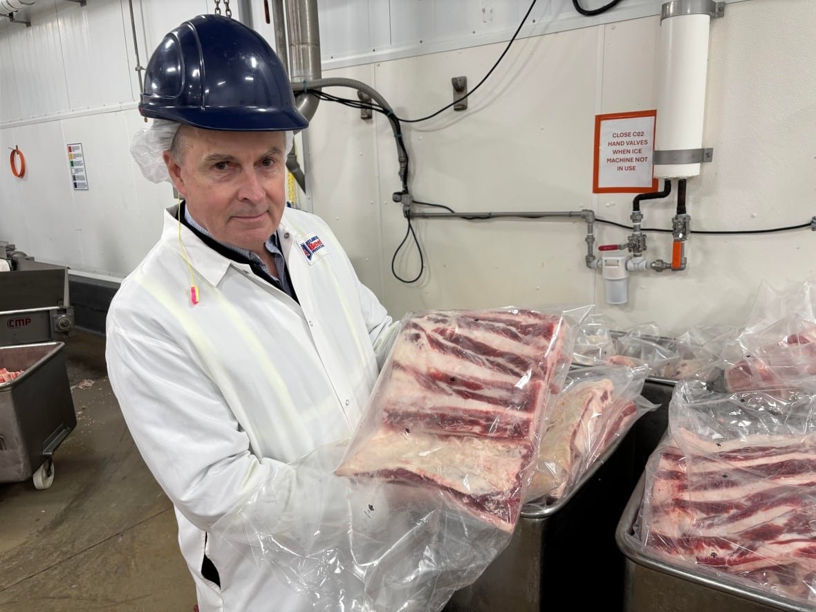 A man in a white lab coat and a dark hard hat holds up a large cut of beef in a plastic bag in a meat processing facility.