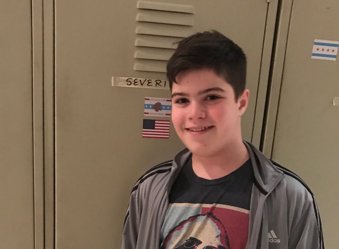 A teenage boy with short, dark hair smiles while standing in front of a school locker.