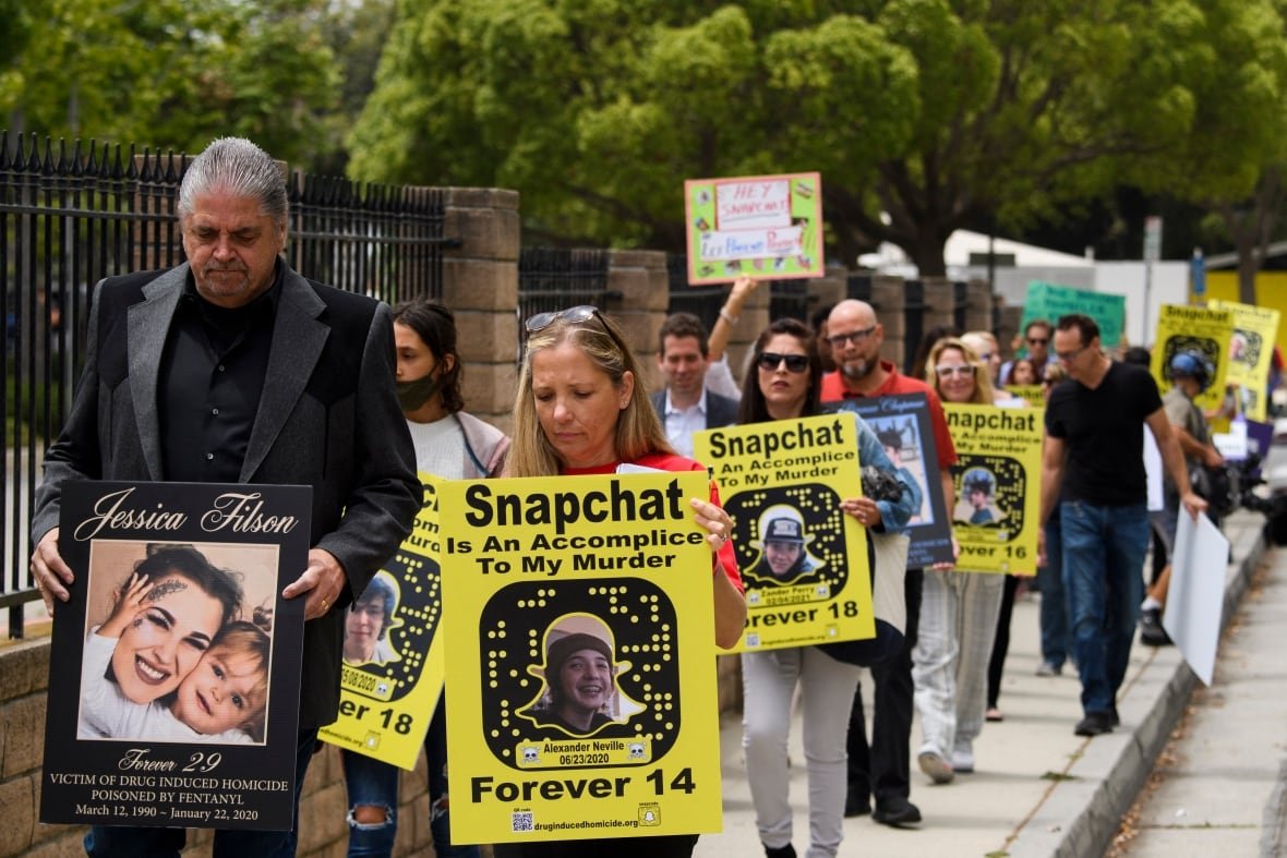 A group of people walk along a sidewalk carrying yellow placards reading 'Snapchat is an accomplice to my murder' with the Snapchat logo and pictures of their loved ones and the age at which they died of drug overdoses.