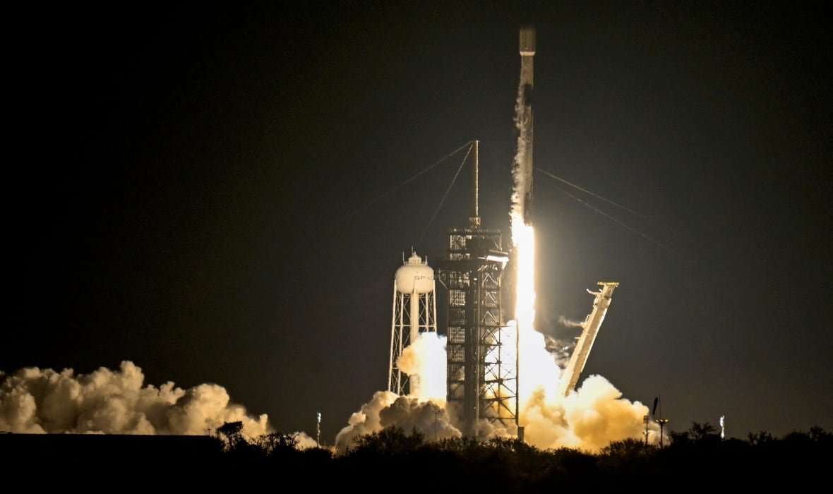 A rocket blasts off from the launchpad, sending orange and white clouds of smoke billowing out from underneath it. It has only just started to move up in this image and is still close to launch tower.