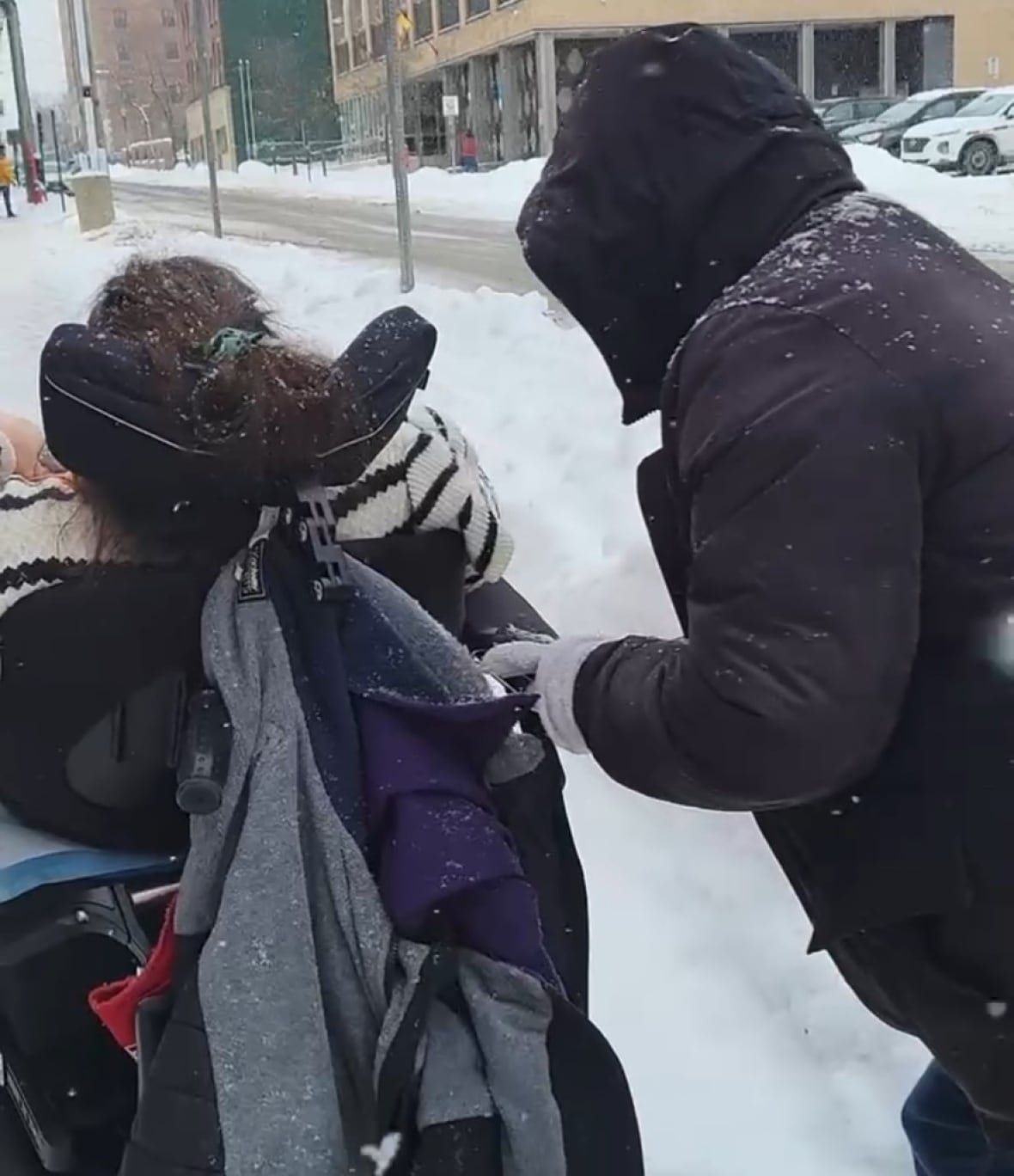 stacey-howe-wheelchair-stuck Seen from behind, a woman in a power wheelchair with a man, wearing a black coat, stands behind her, pushing.