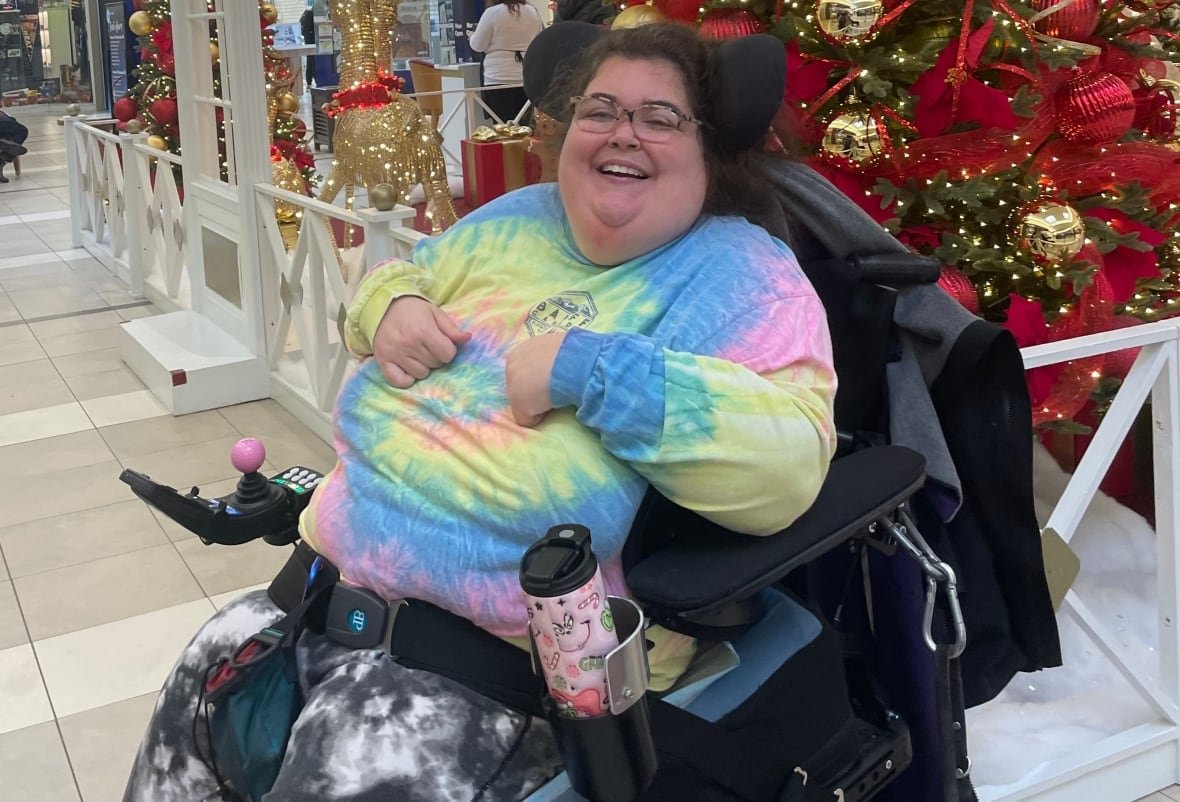 stacey-howe A smiling woman, wearing a tie-dye shirt, sitting in a power wheelchair in front of a Christmas tree covered in red bows.