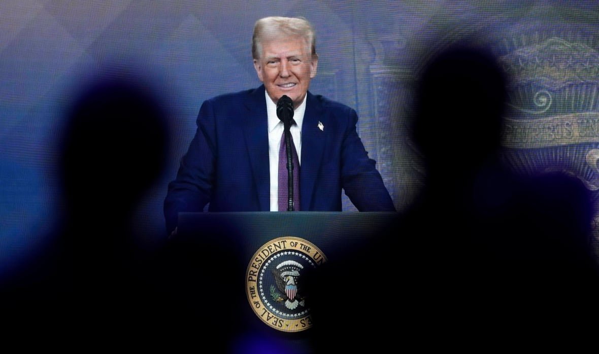 A man standing at a lectern smiles while addressing a crowd. Two people, seen from behind in silhouette, are watching.