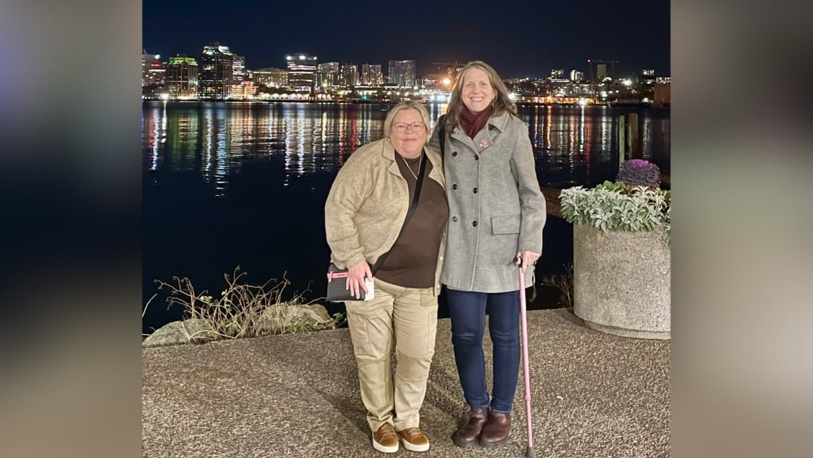 Two women stand in front of a body of water with a city skyline in the background. It is night.