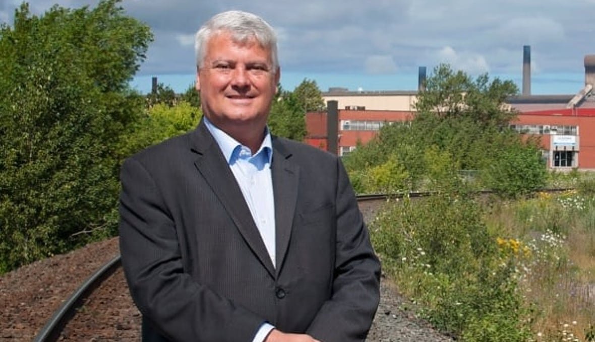 a man in a suit with grey hair stand smiling for a photo with a factory in the background