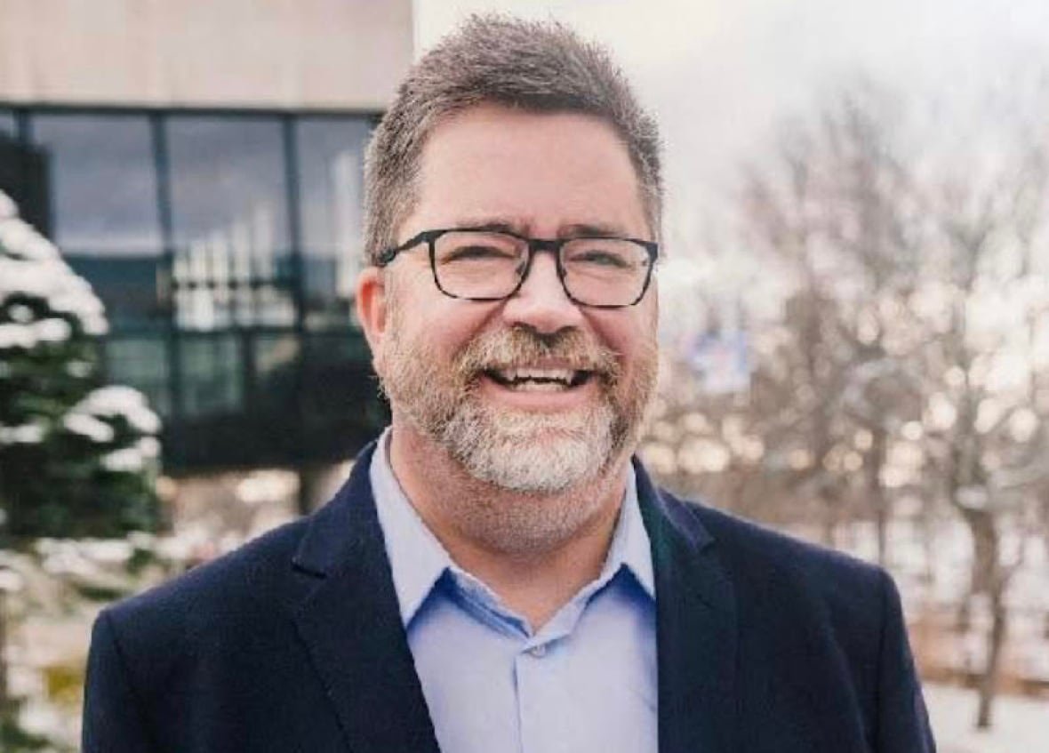 A man with a beard and a black suit jacket smiles at the camera with a building and a bit of snow in the background.