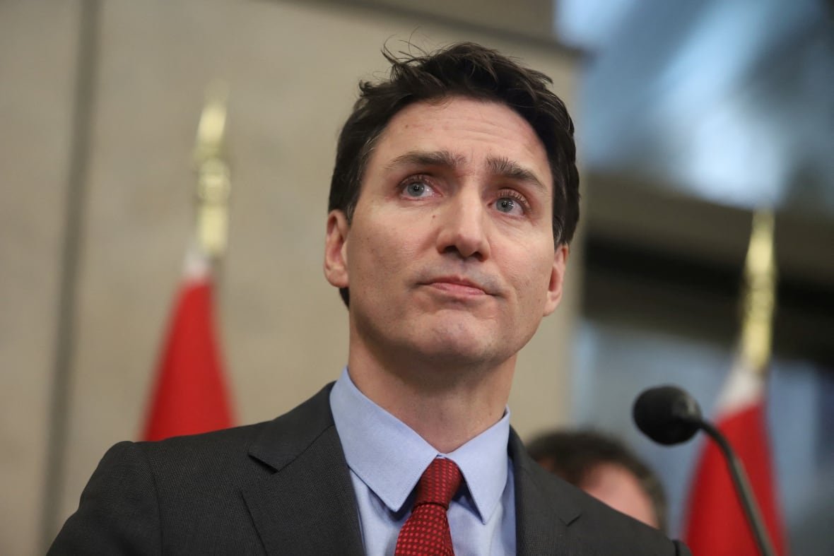 trudeau-tariffs-2 A man in a grey suit, blue shirt and red tie stands at a podium with a stern expression.
