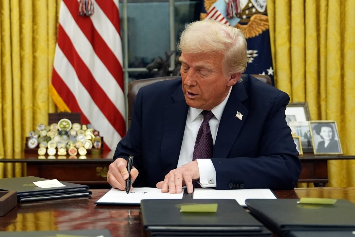 A man sitting at a desk signs documents in one of several black folders stacked around him.